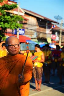 Ein lächelnder glatzköpfiger Mönch, der ein leuchtend oranges Gewand trägt, macht ein Foto von sich und einer Parade im Hintergrund mit einem Selfie-Stick